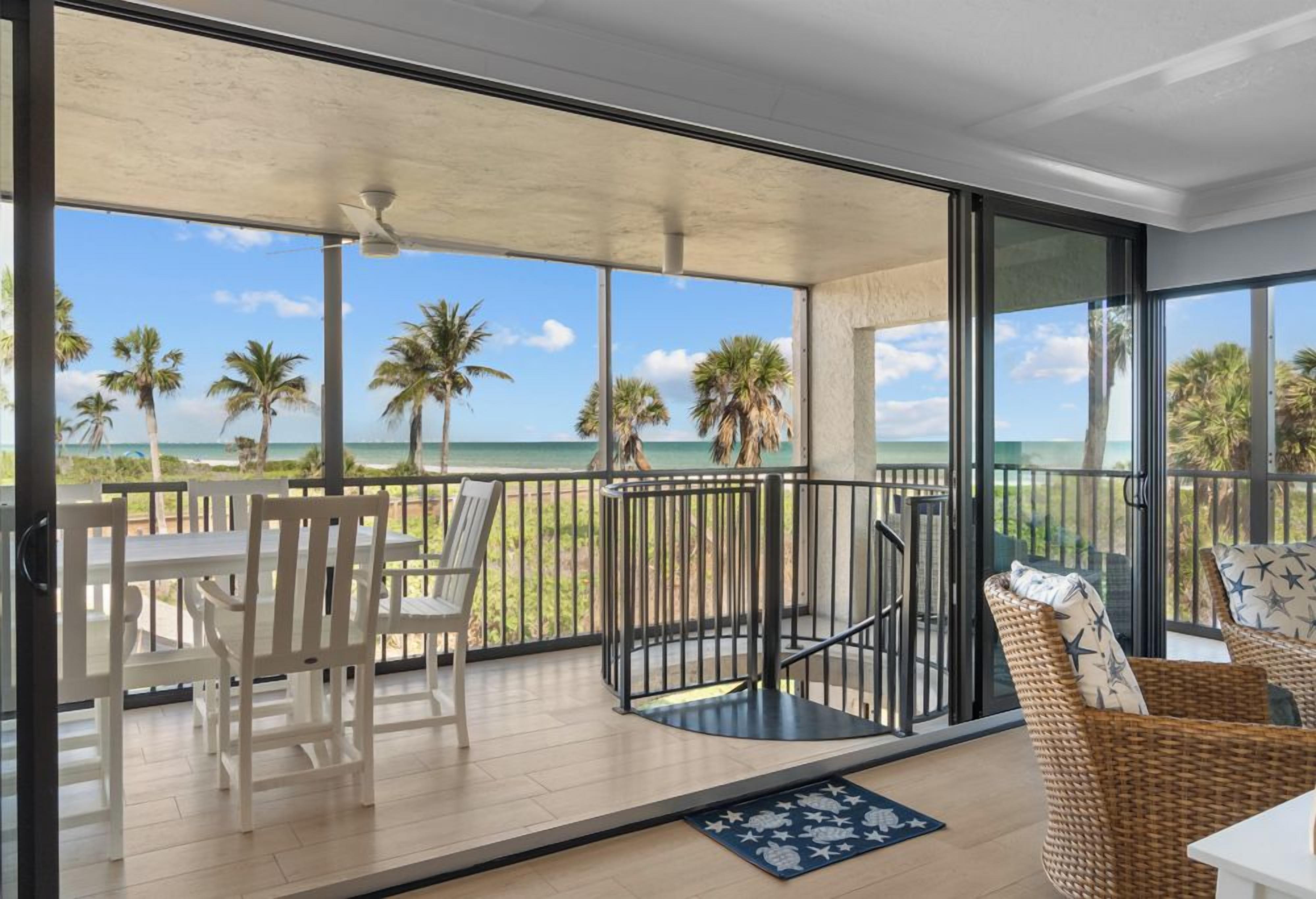 Een balkon met uitzicht op het strand en palmbomen, ingericht met witte stoelen en rieten meubels.