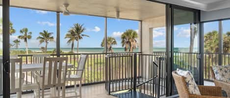 Een balkon met uitzicht op het strand en palmbomen, ingericht met witte stoelen en rieten meubels.