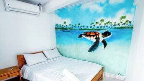 Un dormitorio con cama, una mesita de noche de madera, pared blanca y un mural que muestra una tortuga nadando en el océano con palmeras.