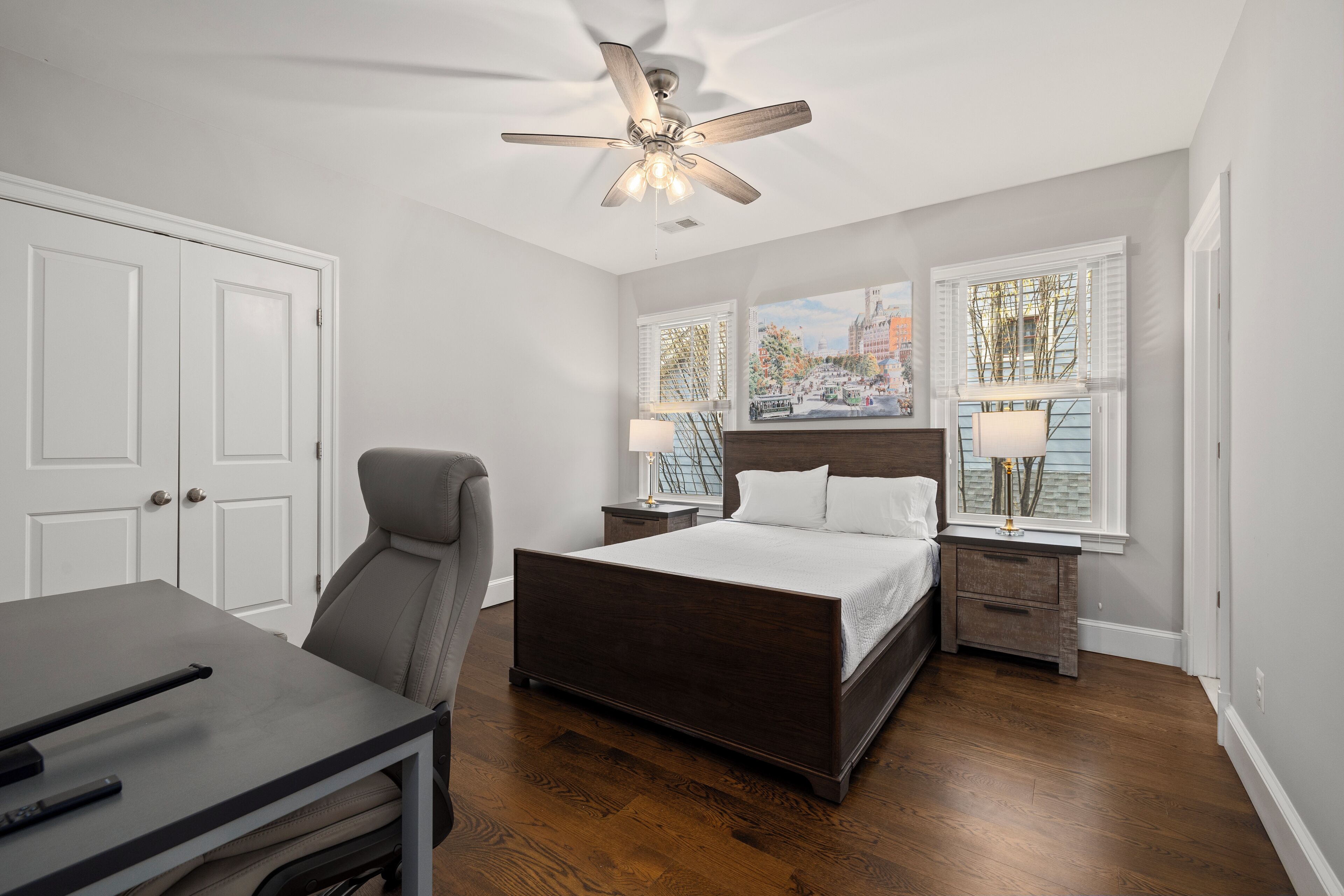 A bedroom with a bed, a desk, a chair, a ceiling fan, and two windows with artwork.
