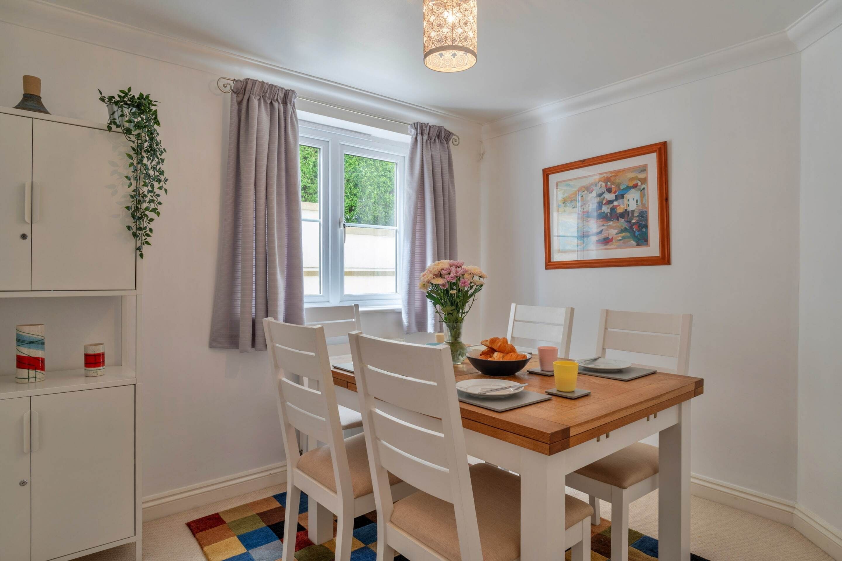 A dining area with a wooden table, white chairs, a vase with flowers, and a painting on the wall.