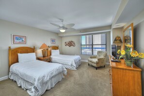 A bedroom with two beds, a ceiling fan, a window with blinds, a chair, a dresser, and a mirror. - Ocean View 3BR-Forest Dunes 1403-Pools & Hot Tub (Myrtle Beach)