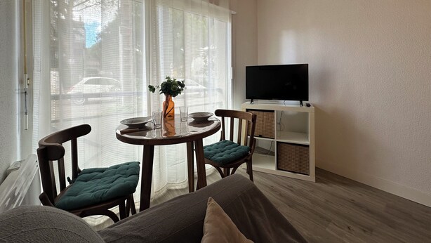 Een woonkamer met een ronde tafel, twee stoelen, een bank, een televisie op een dressoir en een raam met dunne gordijnen.