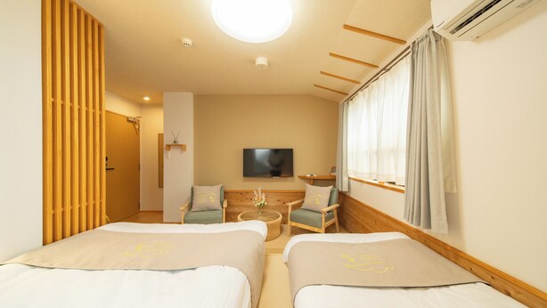 A hotel room with two beds, a wooden partition, a TV, a small table, and a window with curtains.