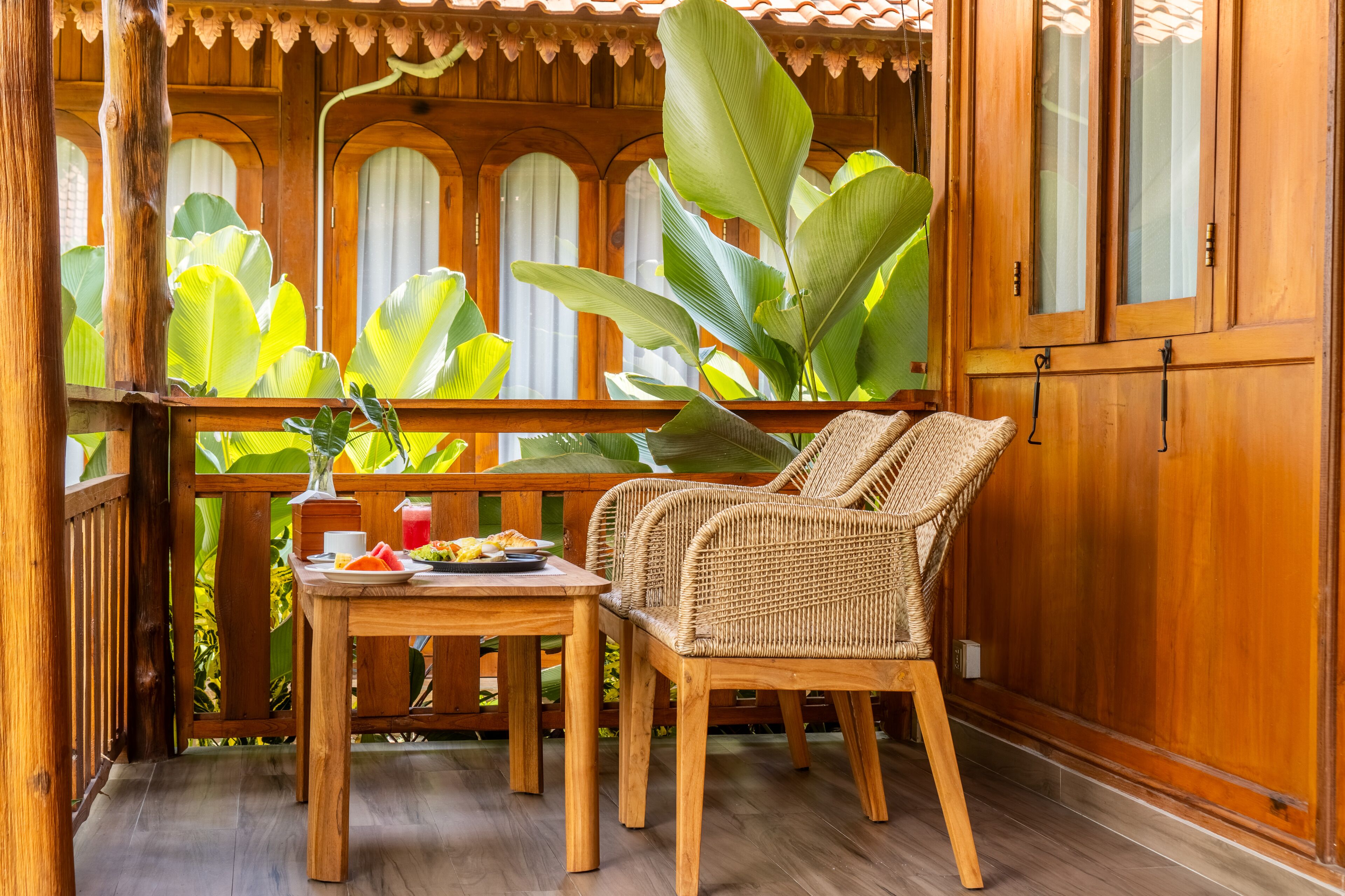A wooden deck with a table and two wicker chairs, surrounded by large green plants and wooden railings.
