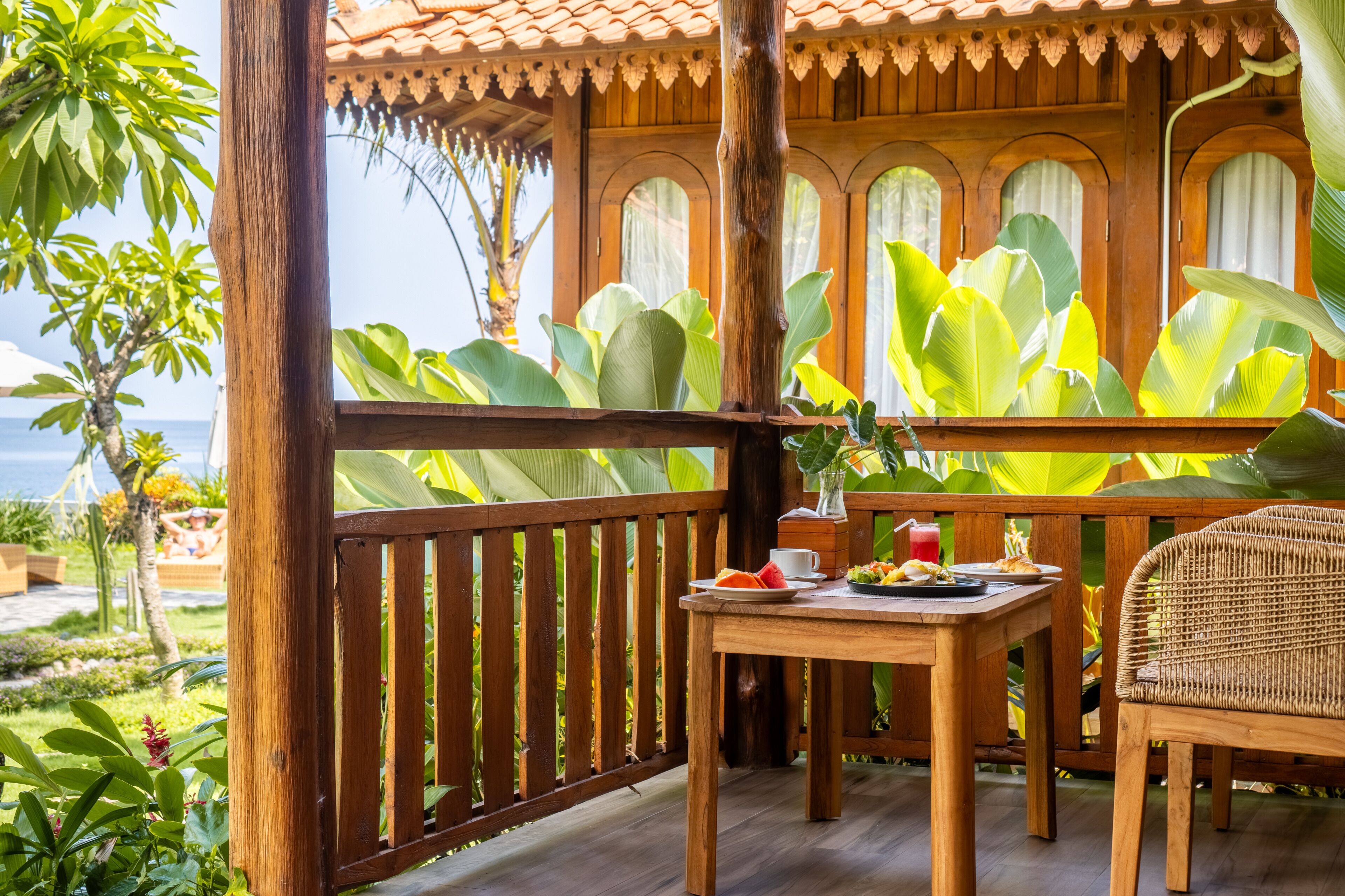 A wooden deck with a table and chairs, surrounded by lush tropical plants and a view of the ocean.