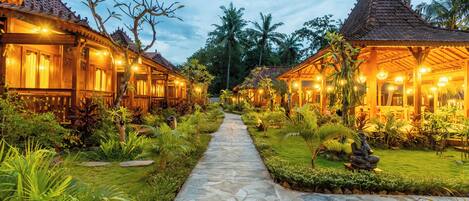 A pathway leading to traditional-style buildings with warm lighting, surrounded by lush greenery and palm trees.