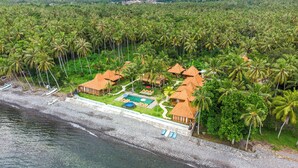 An aerial view of a resort with multiple bungalow-style buildings, a swimming pool, and a beach area with boats. - Dani Beach Villa Penuktukan (Tejakula)