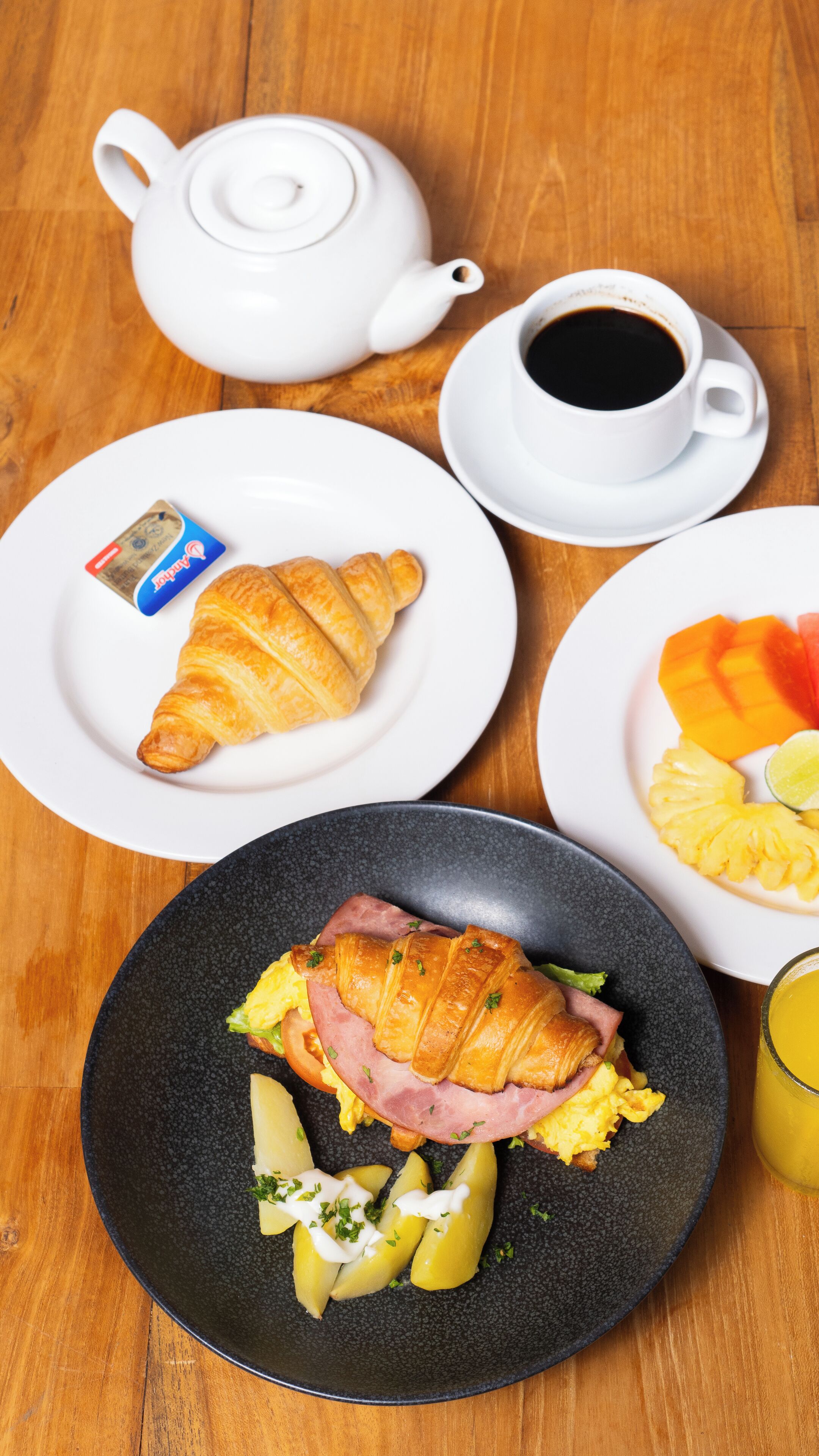 A breakfast setting with croissants, ham, eggs, and fruit on plates, a teapot, a cup of coffee, and a juice glass.