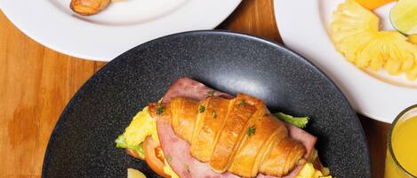 A breakfast setting with croissants, ham, eggs, and fruit on plates, a teapot, a cup of coffee, and a juice glass.