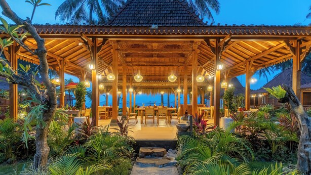 A covered outdoor seating area with tables and chairs, surrounded by lush tropical plants and a stone pathway. - Dani Beach Villa Penuktukan (Tejakula)