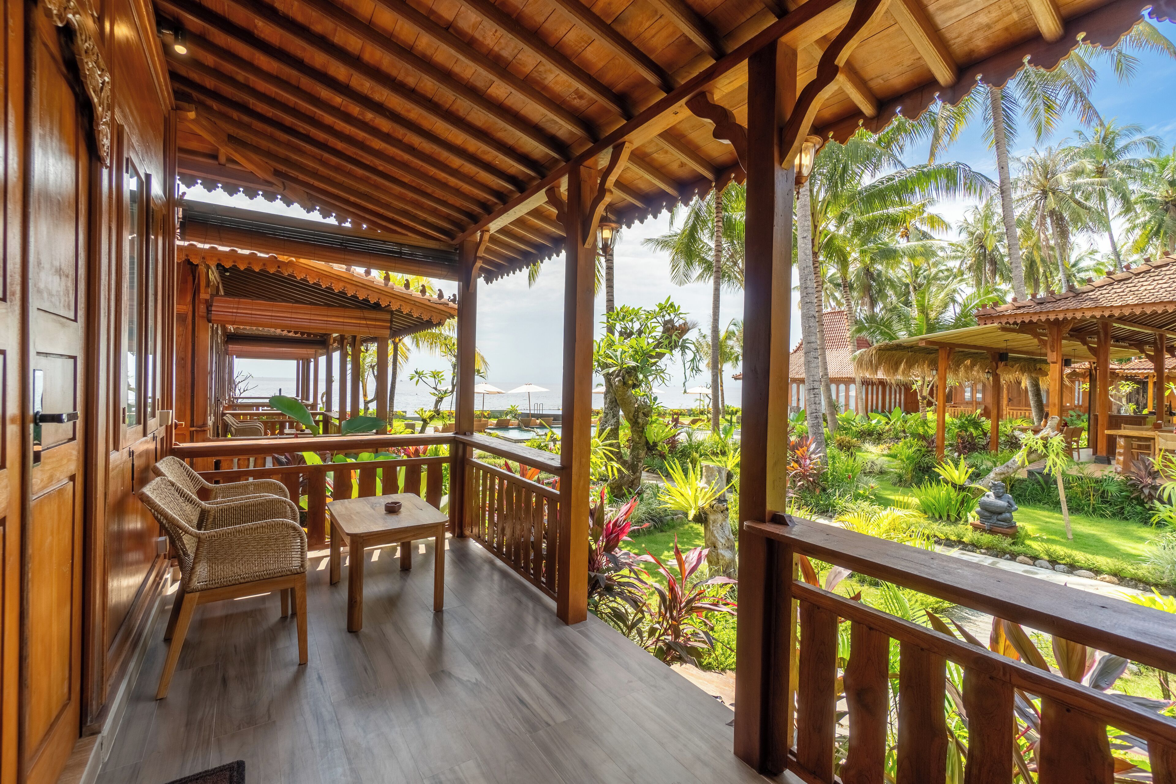 Un balcón de madera con mobiliario de mimbre, vistas a un jardín tropical y una estructura de madera al fondo.