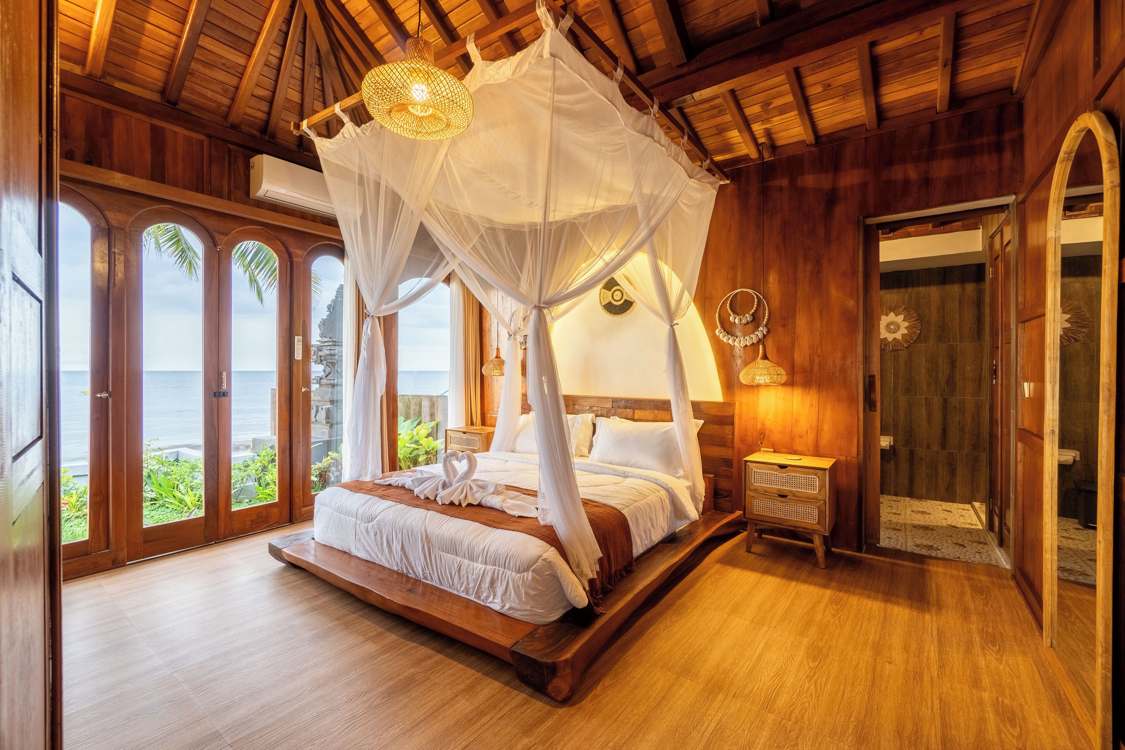 Une chambre à coucher avec un lit à baldaquin, un plancher en bois et une vue sur l’océan grâce à de grandes fenêtres.