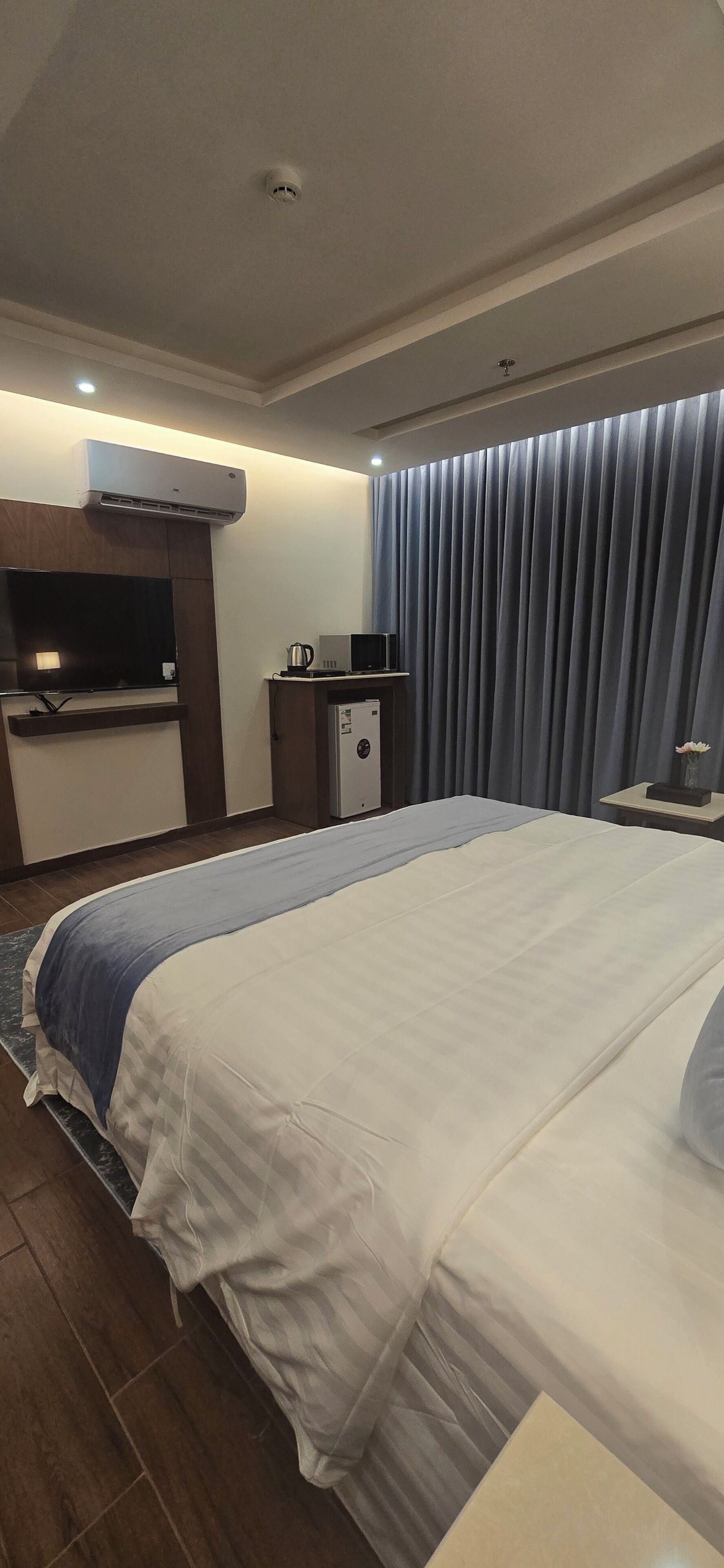 A hotel room with a large bed, a desk with a computer, a television, and an air conditioning unit.