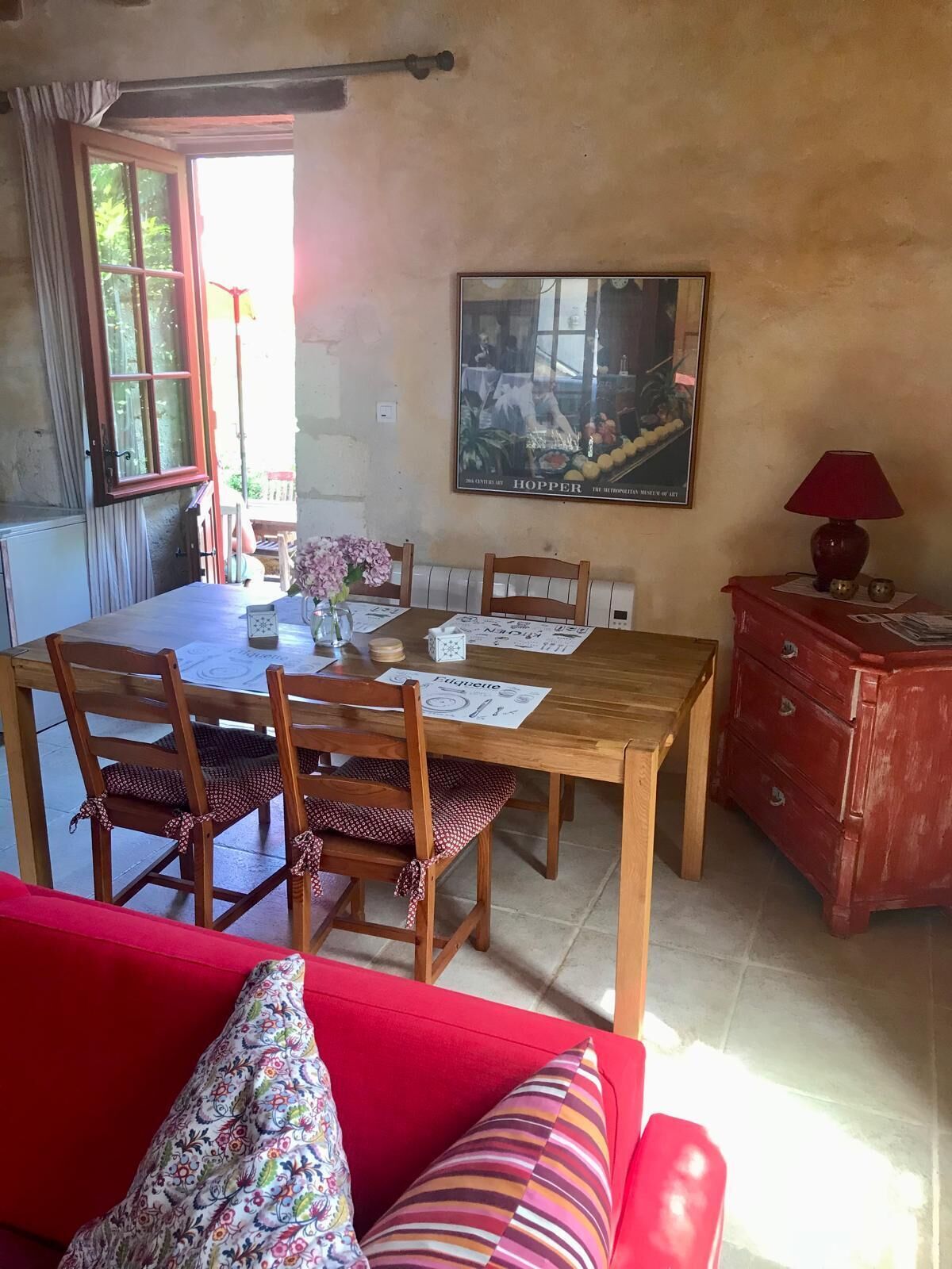 A dining area with a wooden table, chairs, and a red sofa. There is a painting on the wall and a window with curtains.