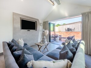 Ein Wohnzimmer mit einer großen, grauen Couch, einem flachbildfernseher an der Wand und einer Glastür, die auf eine Terrasse mit Gartenmöbeln führt.