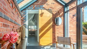 A room with a brick wall, a wooden door, a round mirror, a wicker chair, and a table with flowers. - 1 bedroom accommodation in Meriden, near Coventry (Meriden, near Coventry)