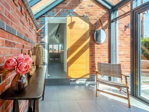 A room with a brick wall, a wooden door, a round mirror, a wicker chair, and a table with flowers. - 1 bedroom accommodation in Meriden, near Coventry (Meriden, near Coventry)