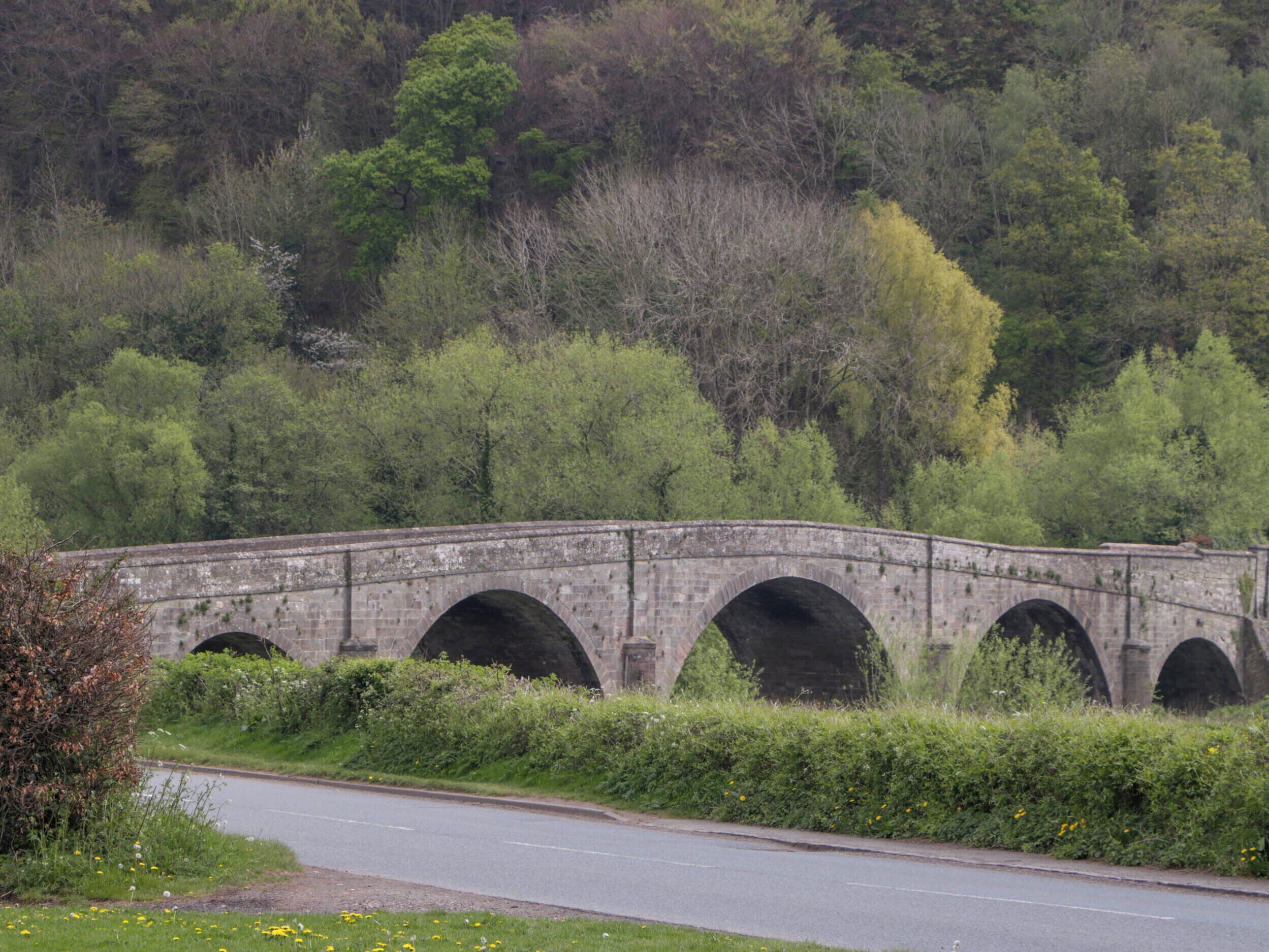 A stone bridge with multiple arches over a road, surrounded by greenery and trees.