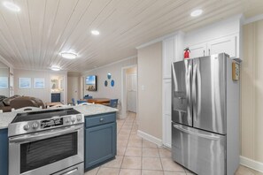 A modern kitchen with stainless steel appliances, a dining area with blue chairs, and a living room with a brown sofa. - 5 BR Dog Friendly Home- Slps 12- Steps from sand (North Myrtle Beach)