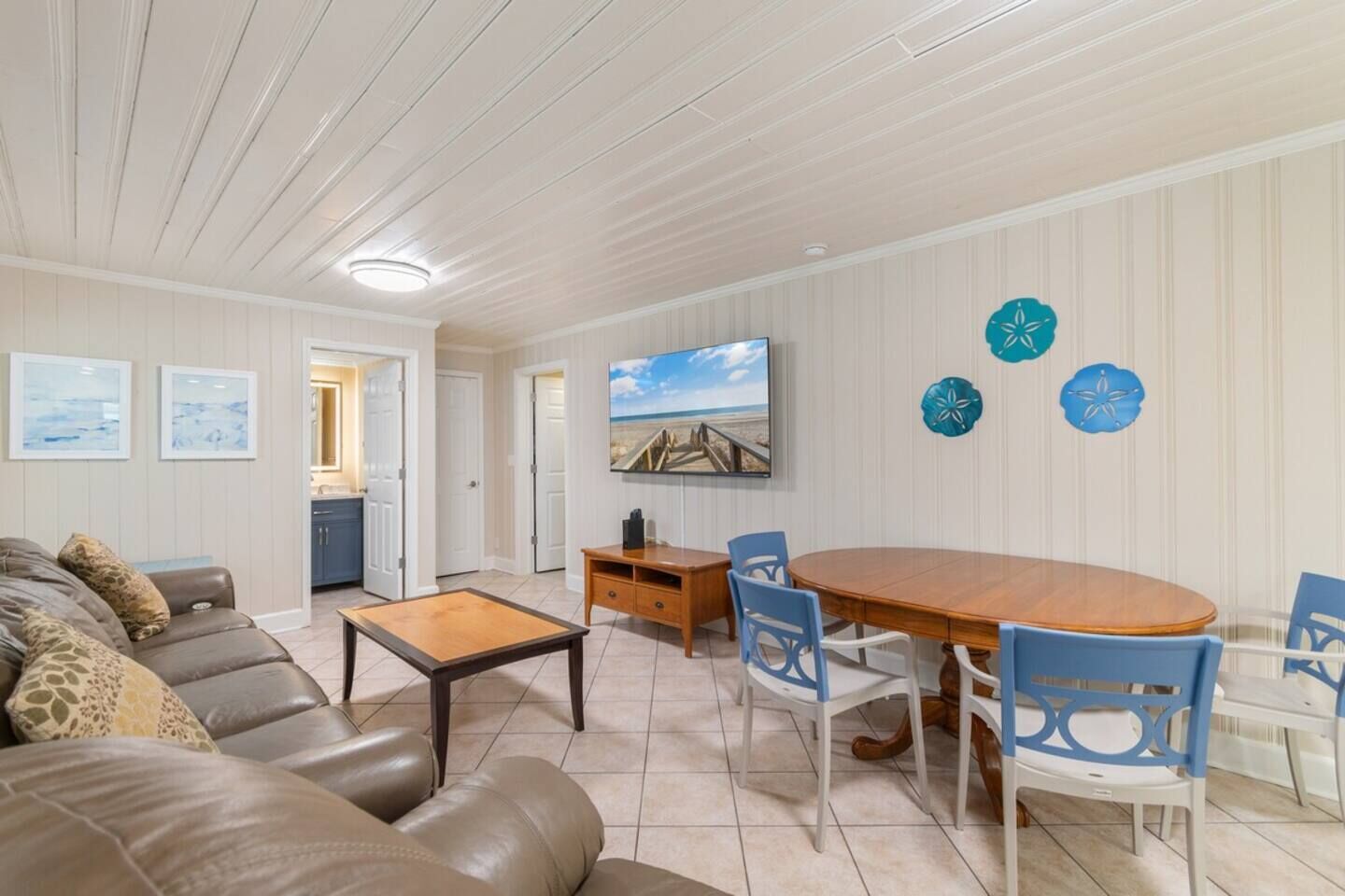 Ett vardagsrum med ett matbord i trä, blå stolar, en soffa i läder och en tv som är monterad på väggen.