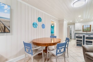 A dining area with a round wooden table and four chairs, a kitchen with white cabinets, and a view of the beach through a window. - 5 BR Dog Friendly Home- Slps 12- Steps from sand (North Myrtle Beach)