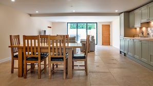 A dining area with wooden chairs and a table, a kitchen with green cabinets, and a sliding glass door leading to an outdoor area. - Barn in Cotswolds With Hot Tub & Scenic Views (Cheltenham)
