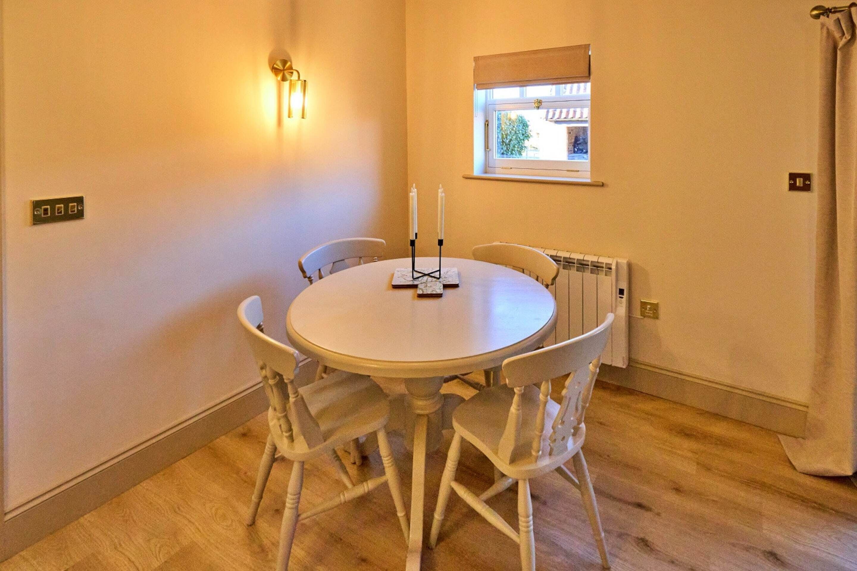 Une table de salle à manger ronde, accompagnée de quatre chaises, un luminaire fixé au mur, une fenêtre donnant sur un paysage agréable et un radiateur.