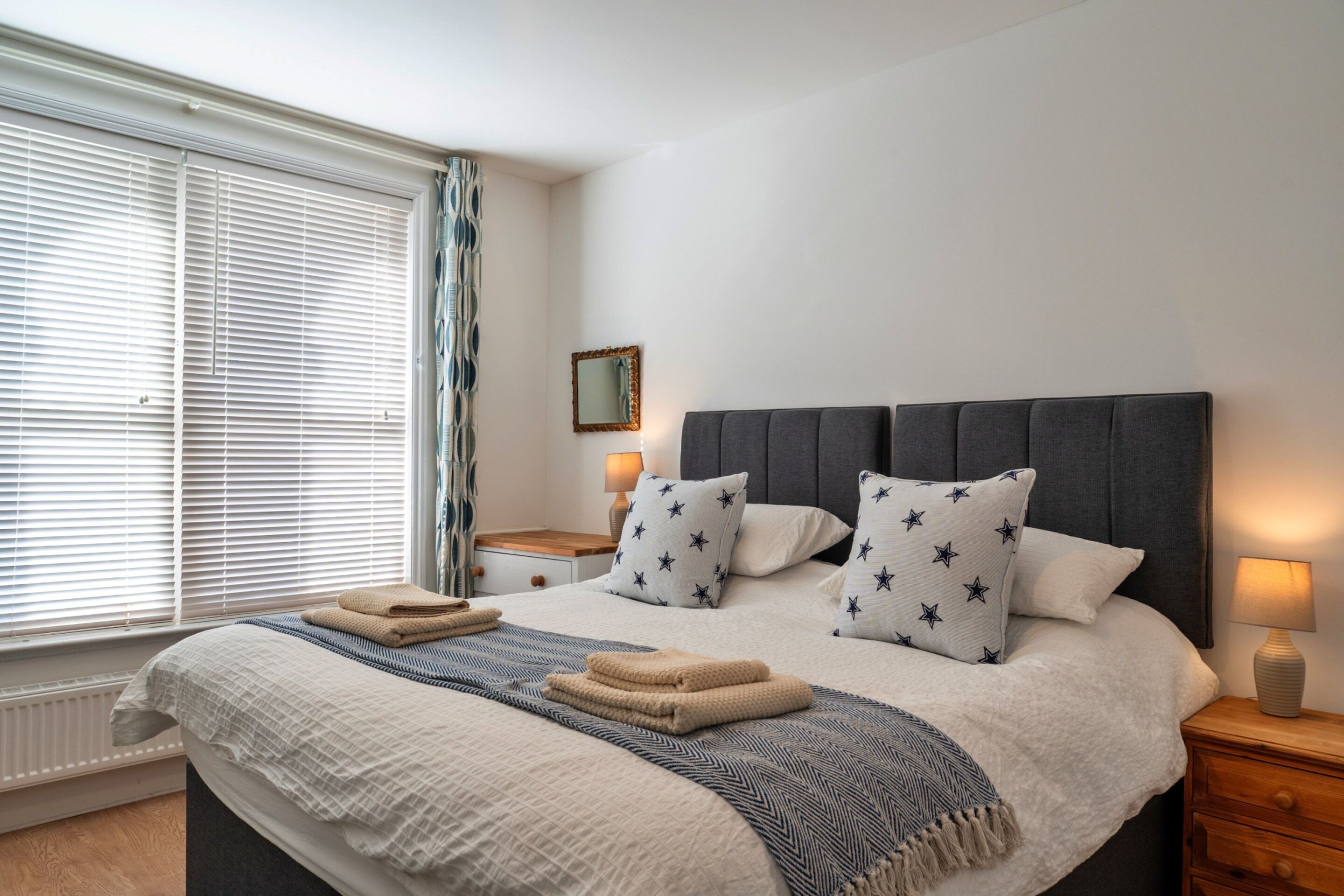 A neatly made bed with star-patterned pillows, a bedside table with a lamp, and a window with blinds.
