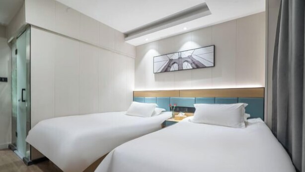 Ein Hotelzimmer mit zwei Betten, einer Glastrennwand, einem hölzernen Kopfteil, einem Wandbild und einem Nachttisch mit Lampe.