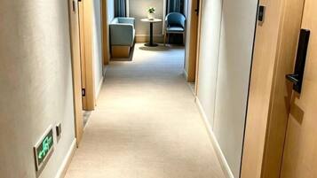 Un couloir d’hôtel avec un sol recouvert de moquette, des portes en bois et un coin salon situé au fond.
