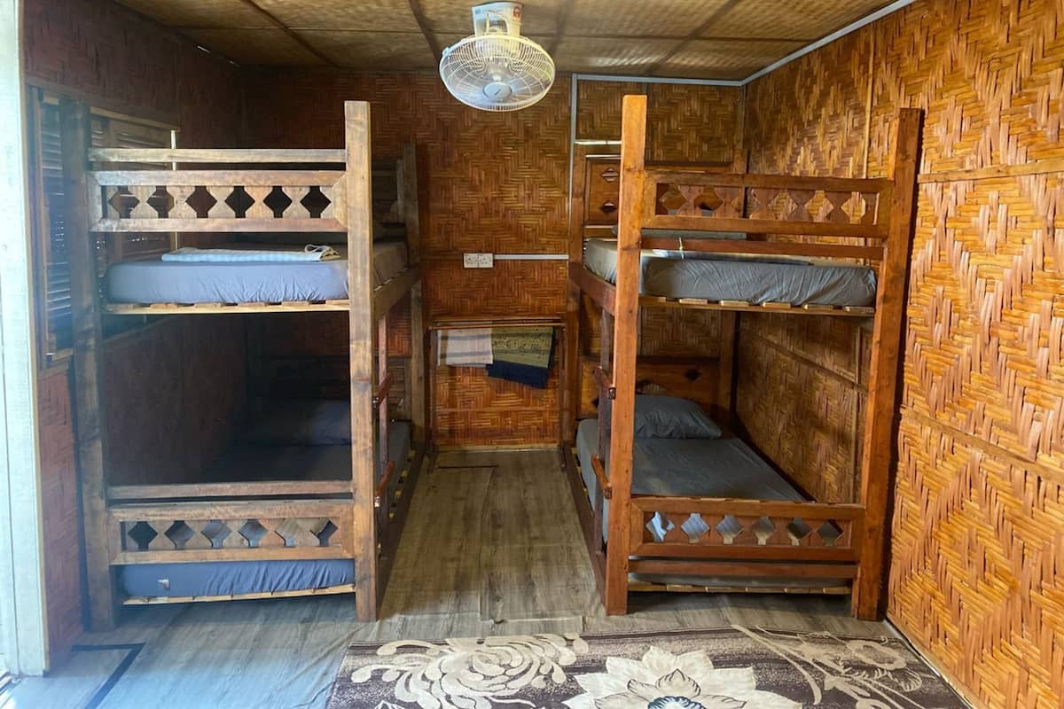 Quarto pequeno e rústico com beliches de madeira, parede de tecido e ventoinha no teto.