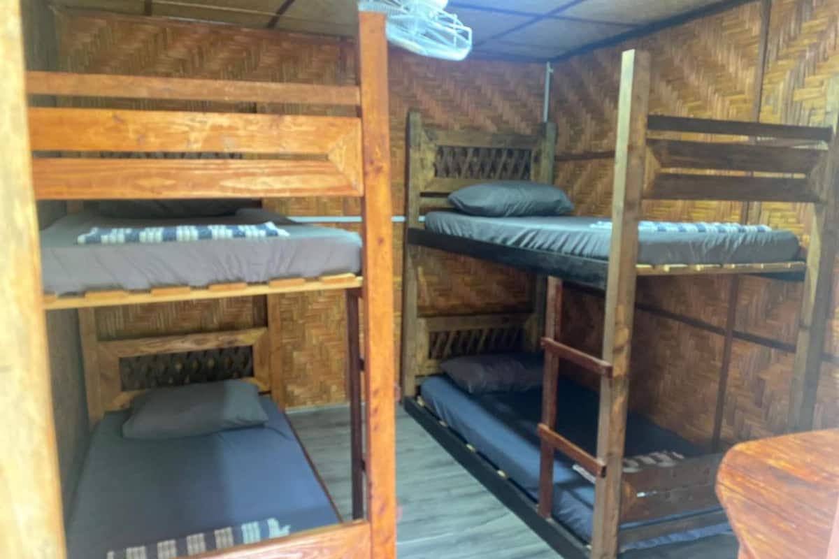 Quarto com beliches, paredes e chão de madeira, uma cama de solteiro com colchão e travesseiro, e uma cadeira de madeira.