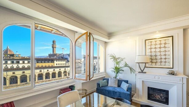 Ein geräumiges Wohnzimmer mit Glastisch, blauem Sofa und Kamin. Große Fenster bieten Blick auf historische Gebäude und einen Turm.