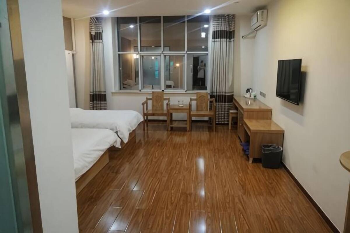A hotel room with a wooden floor, two beds, a desk with a chair, a TV, and a window with curtains.