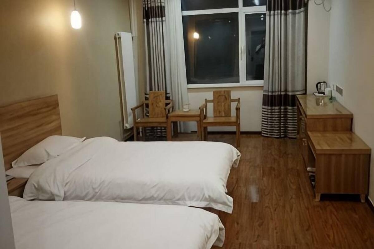 A hotel room with two beds, a wooden desk, a chair, a small table, and a window with curtains.