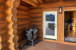 A log cabin with a built-in grill and a door leading to a deck with a view of snow-covered mountains.