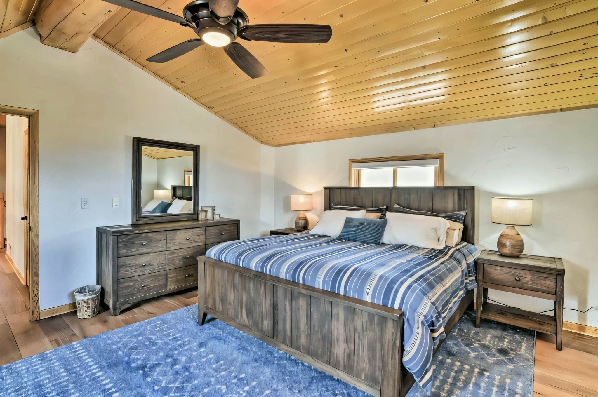 Quarto com teto de madeira, cama com colcha listrada, cômoda, espelho e mesa de cabeceira com abajur.