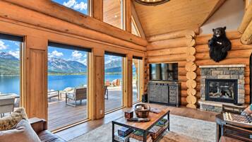 Interior de una cabaña de troncos con chimenea, televisor y vistas a un lago y montañas.