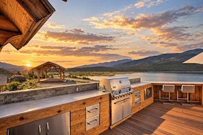 Cocina exterior con parrilla, asientos de bar y vistas a un lago y montañas.