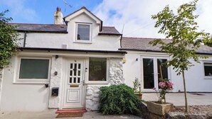 A two-story house with a white exterior, a front door with a small porch, and a stone wall. - Cartref (Holywell)