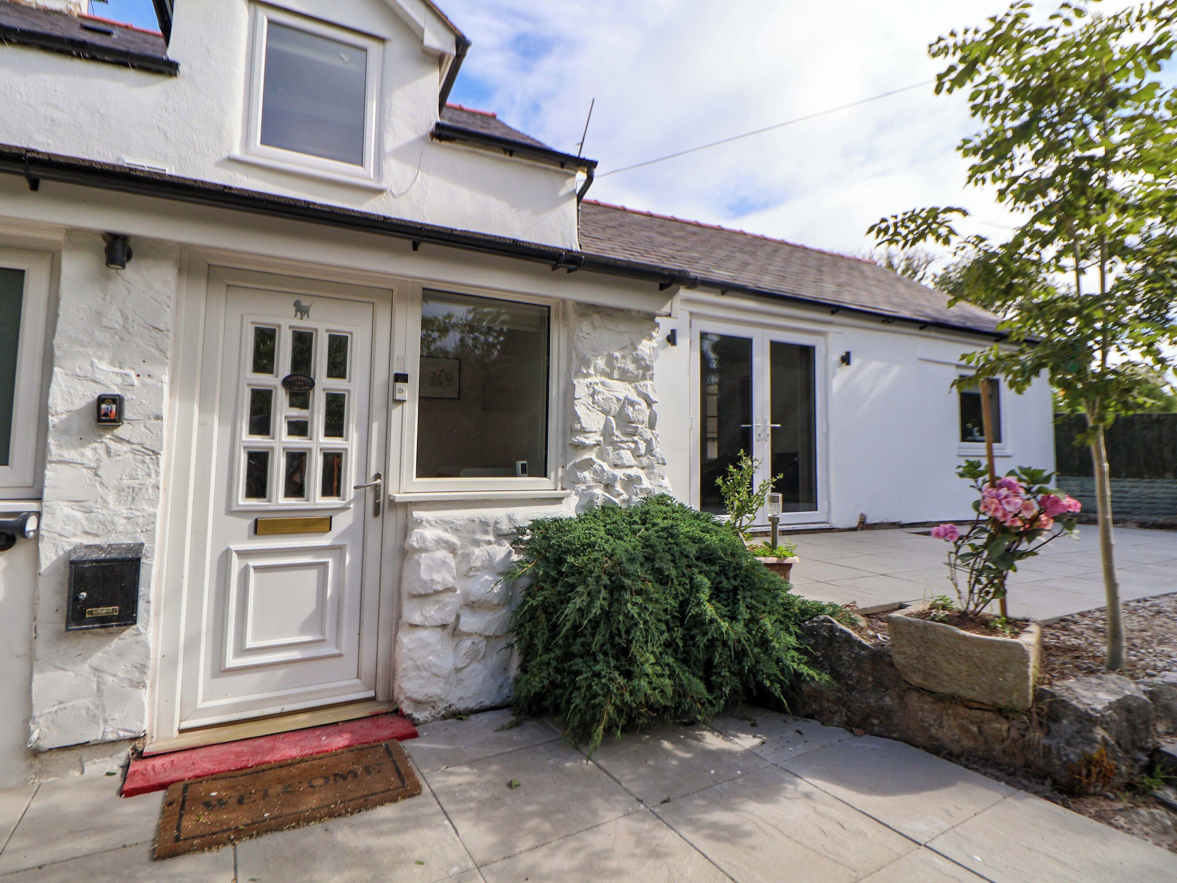 A white building with a stone facade, a red door, and a small garden with pink flowers.
