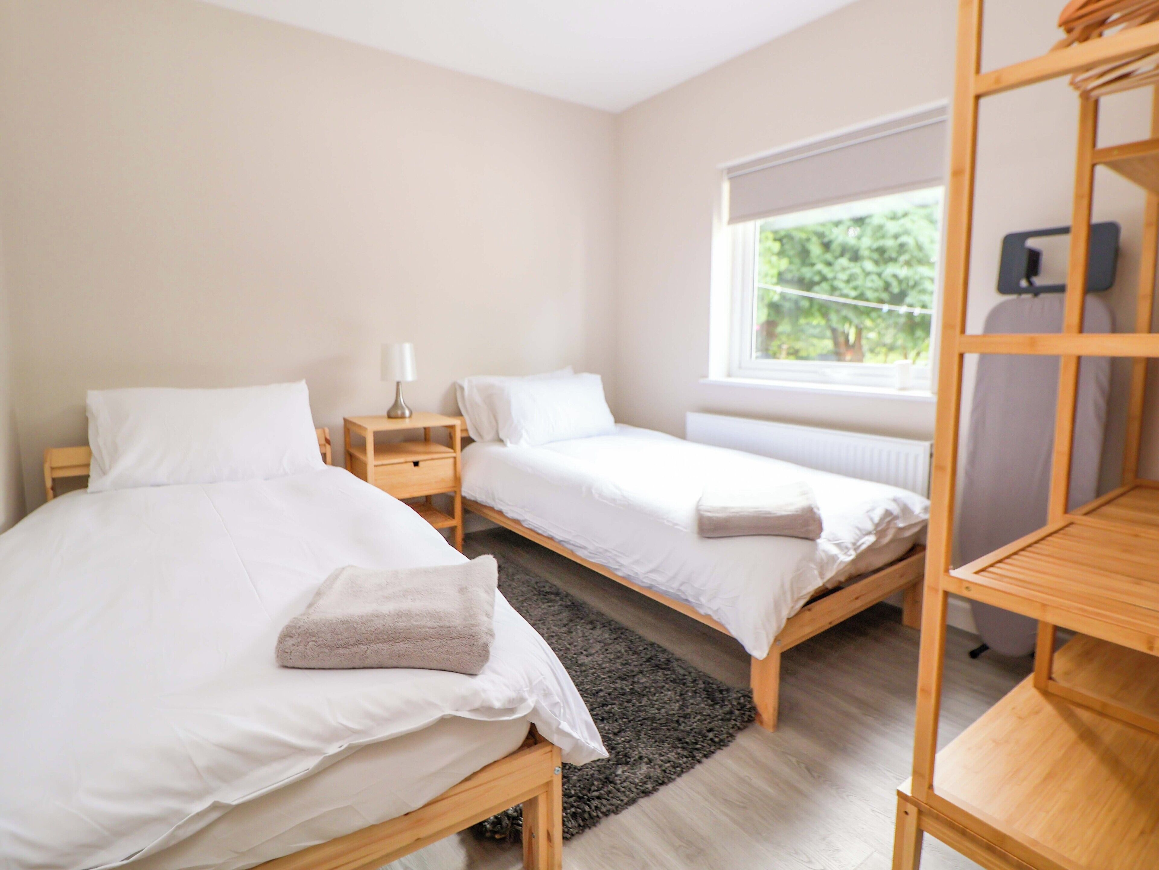 A bedroom with two single beds, a wooden nightstand with a lamp, a window with a view of greenery, and a wooden shelf unit.