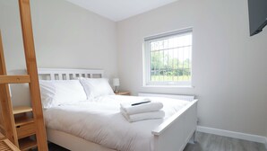 A neatly made bed with white linens and a folded towel, a wooden ladder, a bedside table with a lamp, and a window with a view of greenery. - Cartref (Holywell)