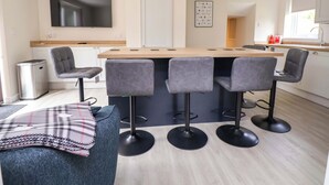 A modern kitchen with a central island, bar stools, and a television mounted on the wall. - Cartref (Holywell)