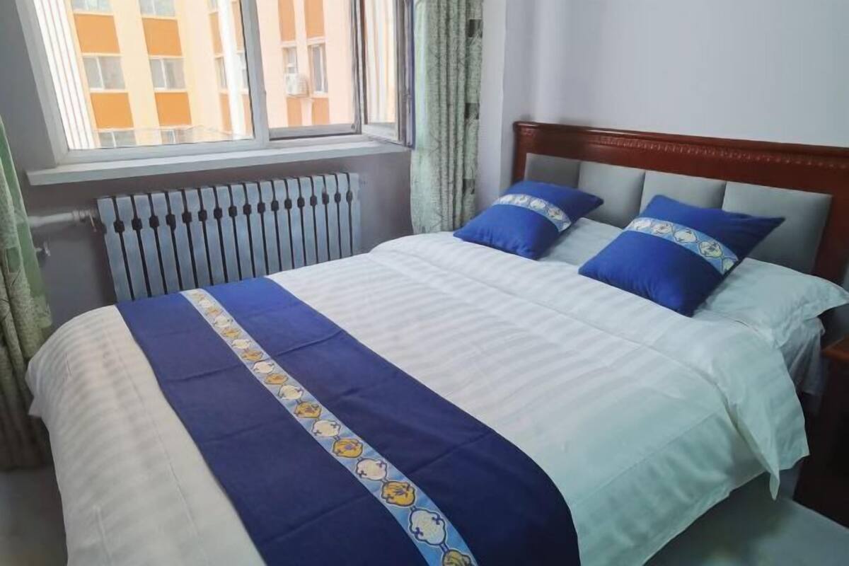 Una cama con una colcha azul y blanca, dos almohadas azules y una ventana que da a vistas de edificios.