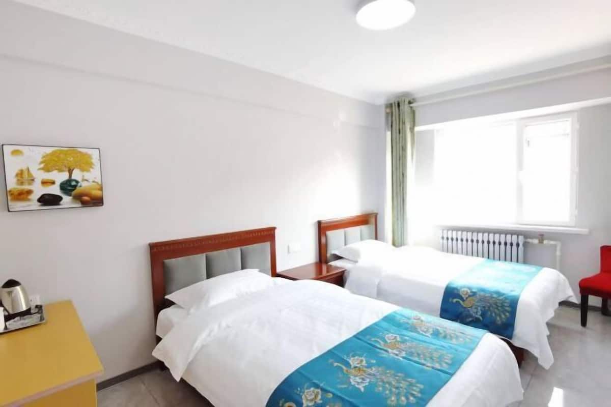 Una habitación de hotel con dos camas, una mesita auxiliar amarilla, una silla roja, una ventana con cortinas y un cuadro en la pared.