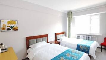 A hotel room with two beds, a yellow side table, a red chair, a window with curtains, and a painting on the wall.