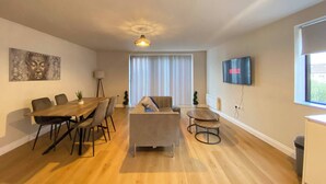 A modern living room with a dining area, a sofa, a TV mounted on the wall, and a window with blinds. - Charming 2-bedroom Apartment in the Heart of Beckenham (Beckenham)