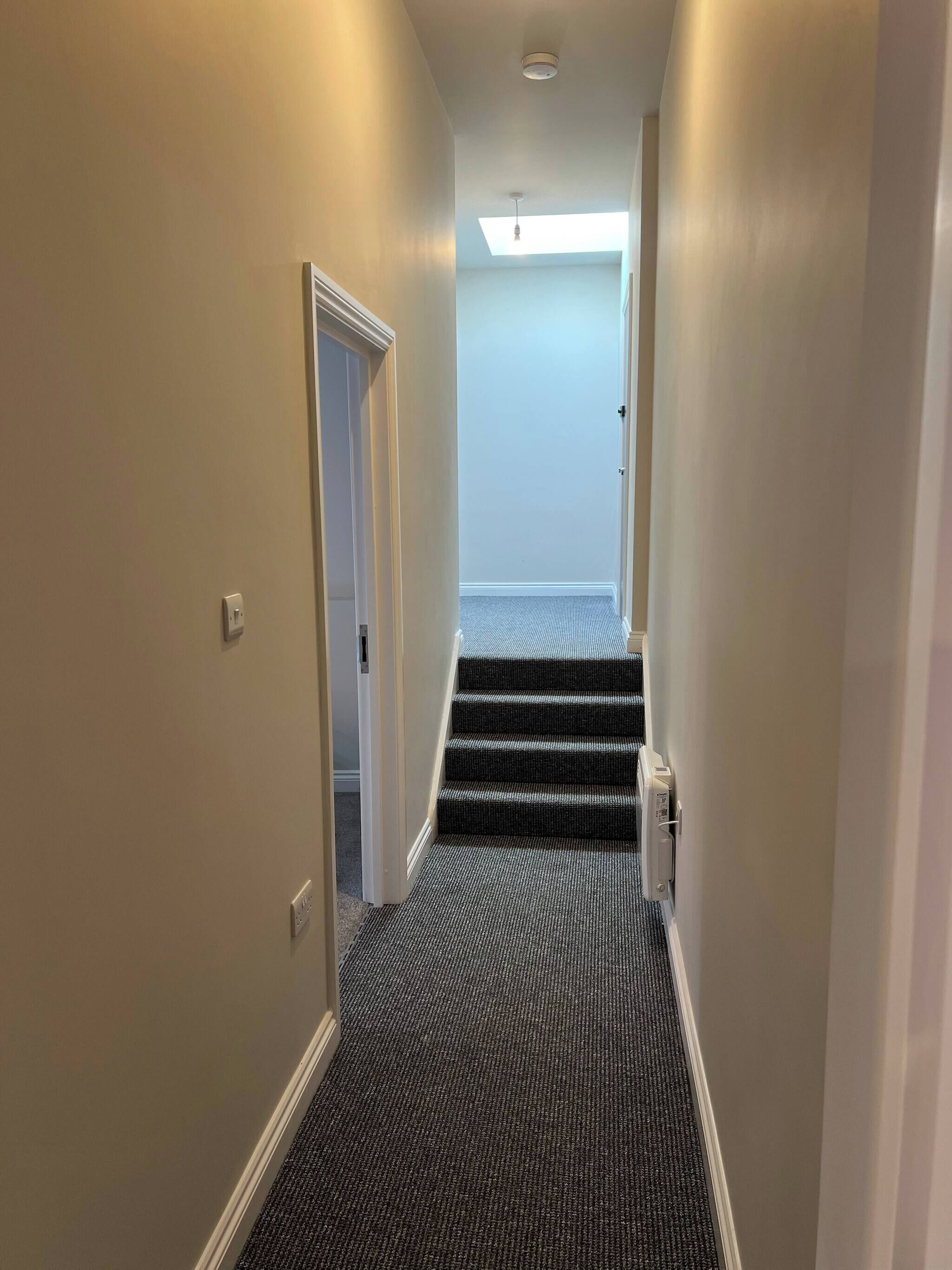 A narrow hallway with carpeted stairs leading to a room with a skylight.